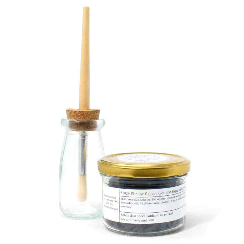 A small glass jar with a gold lid containing dark amber shellac flakes, next to a small clear glass bottle with a cork stopper and a light wooden paint brush, on a white background.