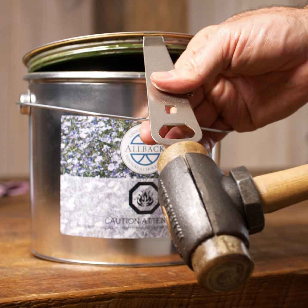Person using the Allbäck Paint Can Opener as a straightedge with hammer to lift the lid on a 3 L metal paint can with rubber gasket.