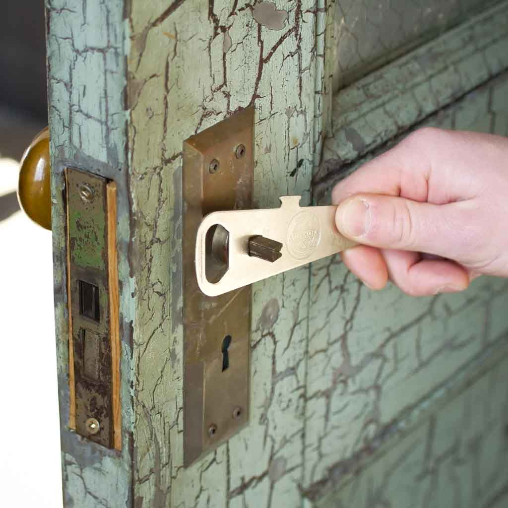 Hand using the Allbäck Paint Can Opener as a temporary door handle to turn the spindle and open an old door.