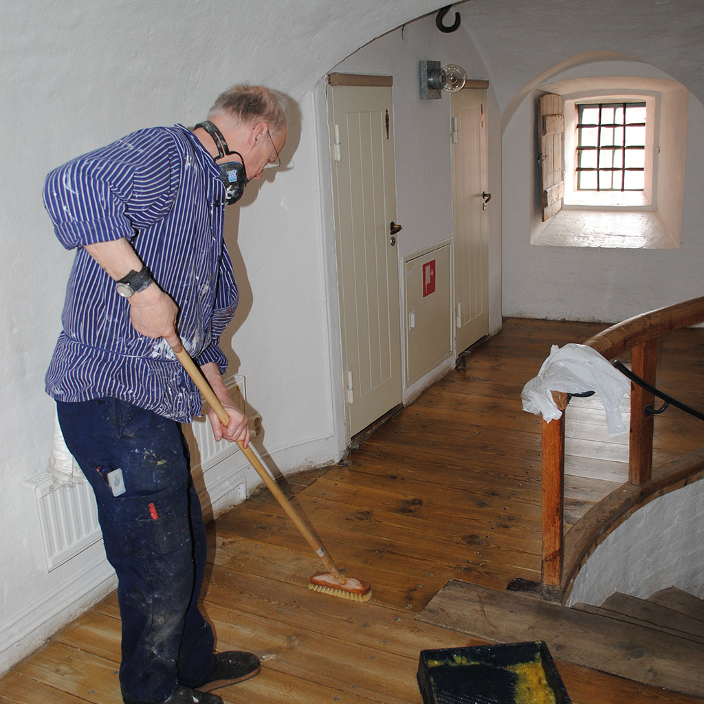 Hans Allbäck applying Linseed Oil Wax to an old pine floor in an old Swedish citadel building, with white plaster walls and a deep-set, old window. From a standing position with a scrub brush attached to an extension pole, Hans is reviving the floor's rich, amber colour tone and its protective sheen.