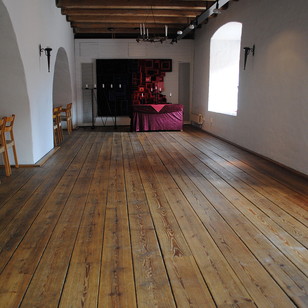 Large room in an old citadel in Sweden, with white plastered walls, a high ceiling with exposed wood beams, and an old pine floor, freshly restored with Allbäck Linseed Oil Wax.