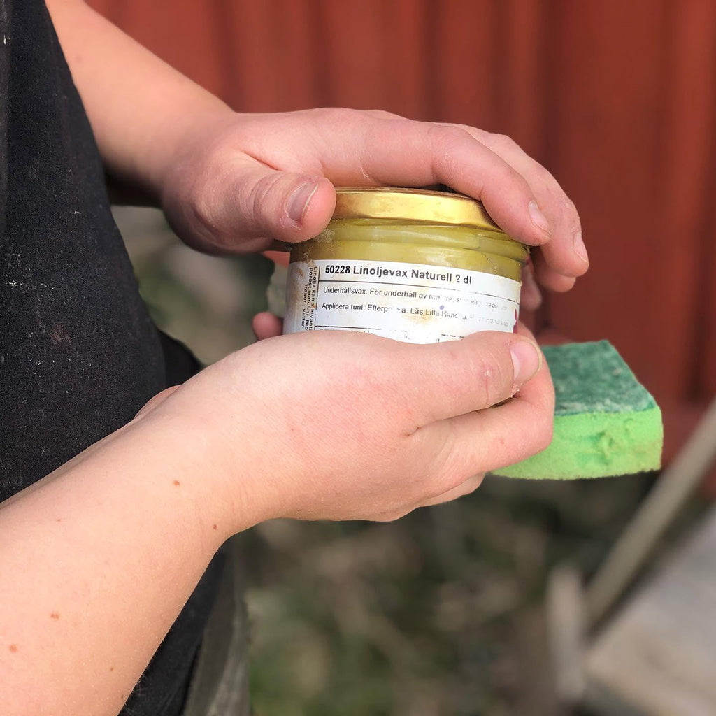 Person holding a jar of Allbäck Linseed Oil Wax Natural (Clear) with a green sponge. Red, painted, vertical wood siding in the background.