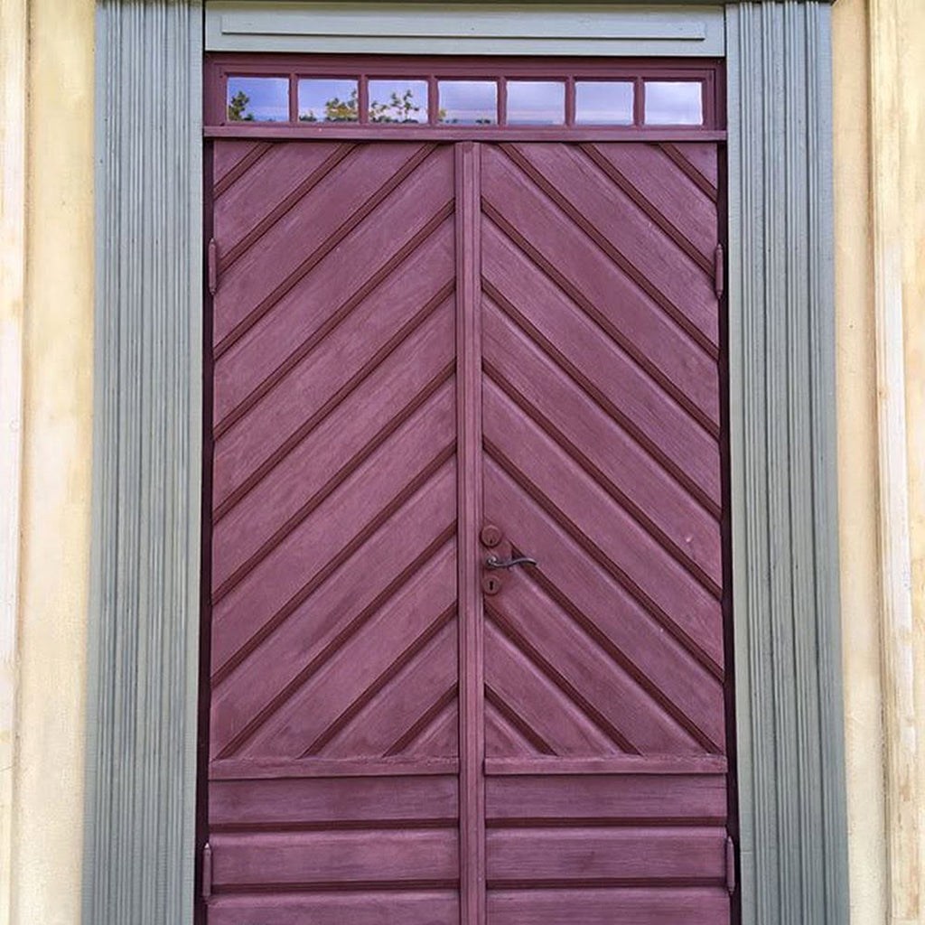 Exterior wooden door with a chevron pattern and glass transom panels, showing faded, chalky red linseed oil paint, ready for maintenance with Allbäck Linseed Oil Wax.