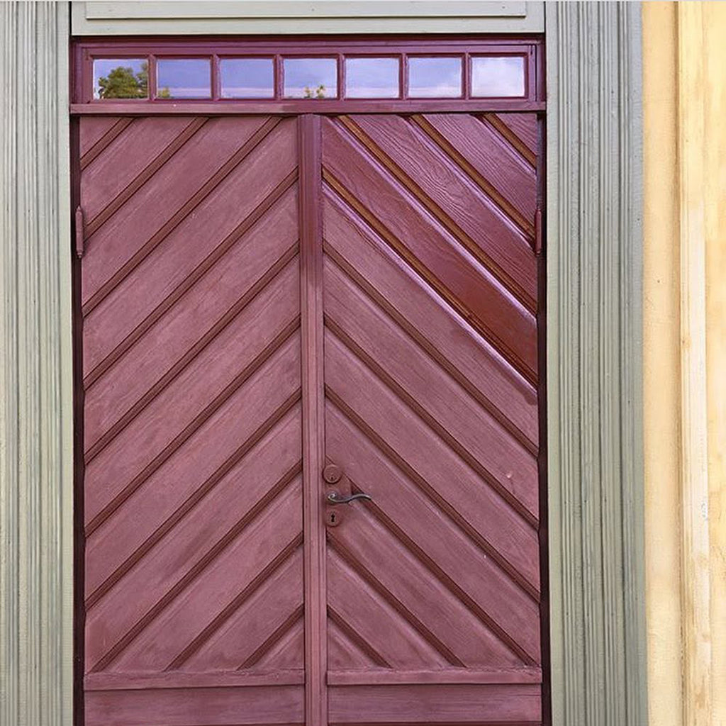 Exterior wooden door with a chevron pattern and glass transom panels, showing faded, chalky red linseed oil paint, partly revived to its original colour and sheen with Allbäck Linseed Oil Wax.