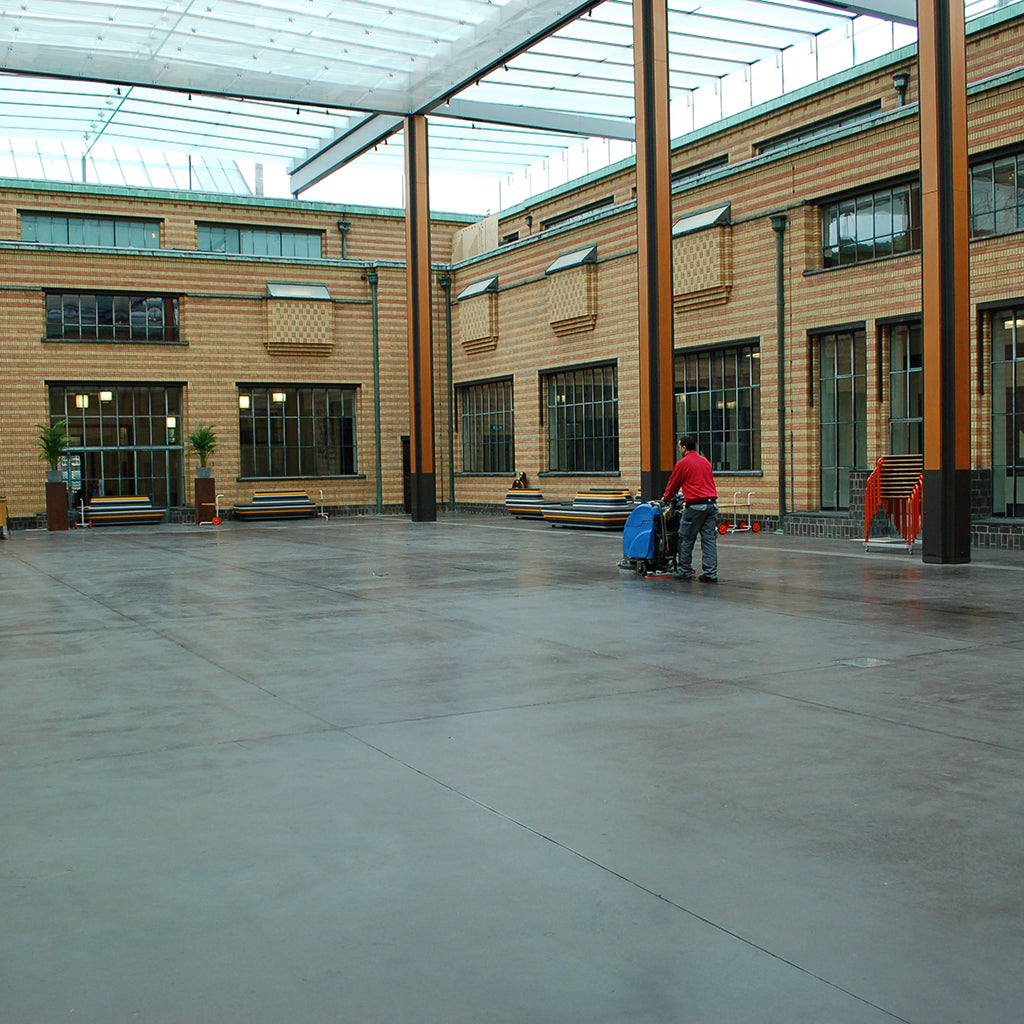 Concrete floor of an inner courtyard, freshly treated with Allbäck Linseed Oil Wax, is being buffed by a man operating a floor buffing machine.