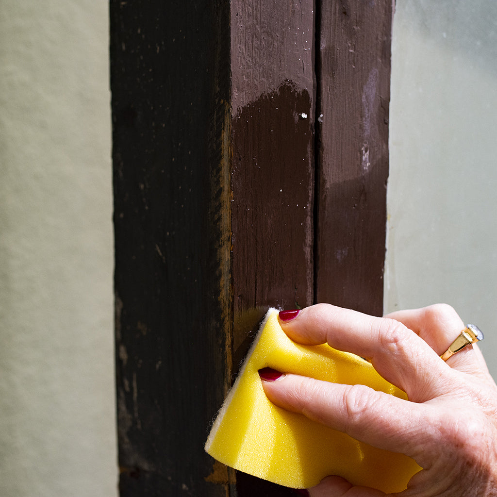 Hand with abrasive yellow sponge applying clear Linseed Oil Wax to revive an old, brown-painted wood surface that has faded over time.
