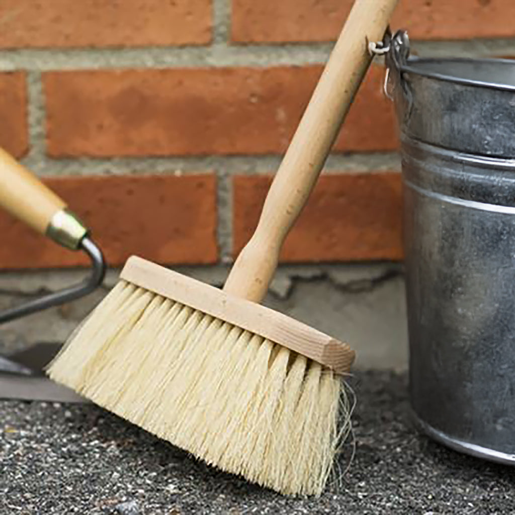Natural Limewash and Masonry Brush leaning against a galvanized metal bucket, with a brick wall and mason's trowel in the background.
