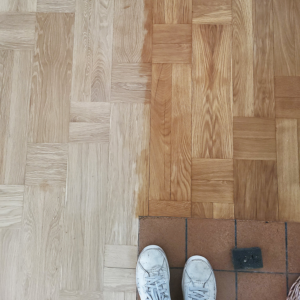 Bare oak, parquet floor, partially treated with Allbäck Linseed Oil Wax, bringing out its rich, amber wood tones. The white shoes of the photographer and a small, black applicator sponge are shown at the bottom of the image for perspective.